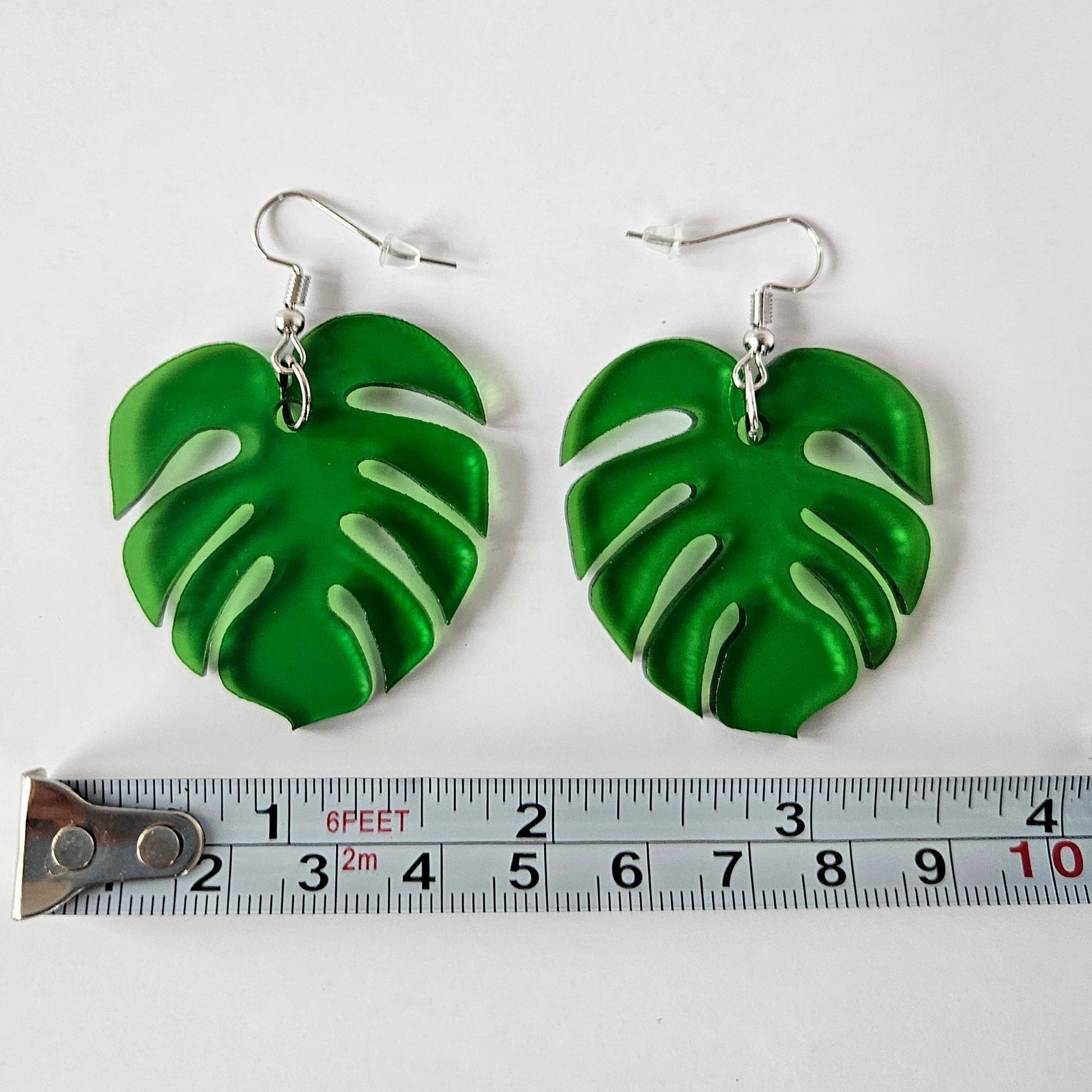 Green Monstera leaf-shaped earrings with a ruler for scale on a white background